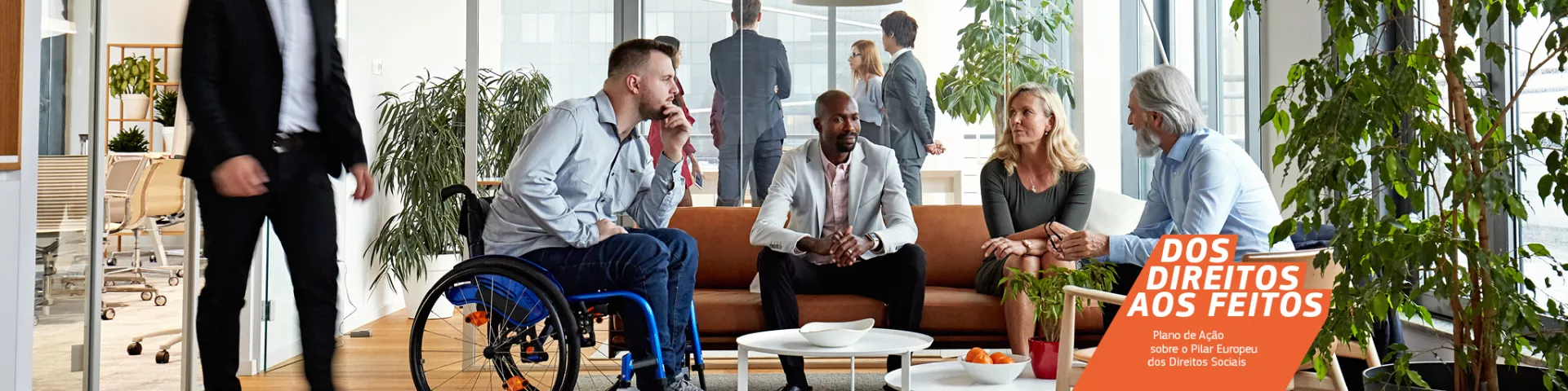 Um grupo diversificado de pessoas, composto por diferentes géneros, idades e etnias, juntos à volta de uma mesa de café baixa num espaço de escritório. Estão a conversar, sentados confortavelmente num sofá e numa cadeira de rodas.