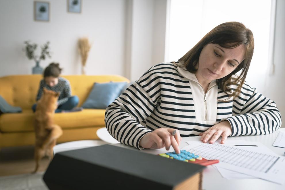 Woman seated at a table using a calculator to process bills. A young child is sitting on a sofa and playing with a dog in the backgroung.
