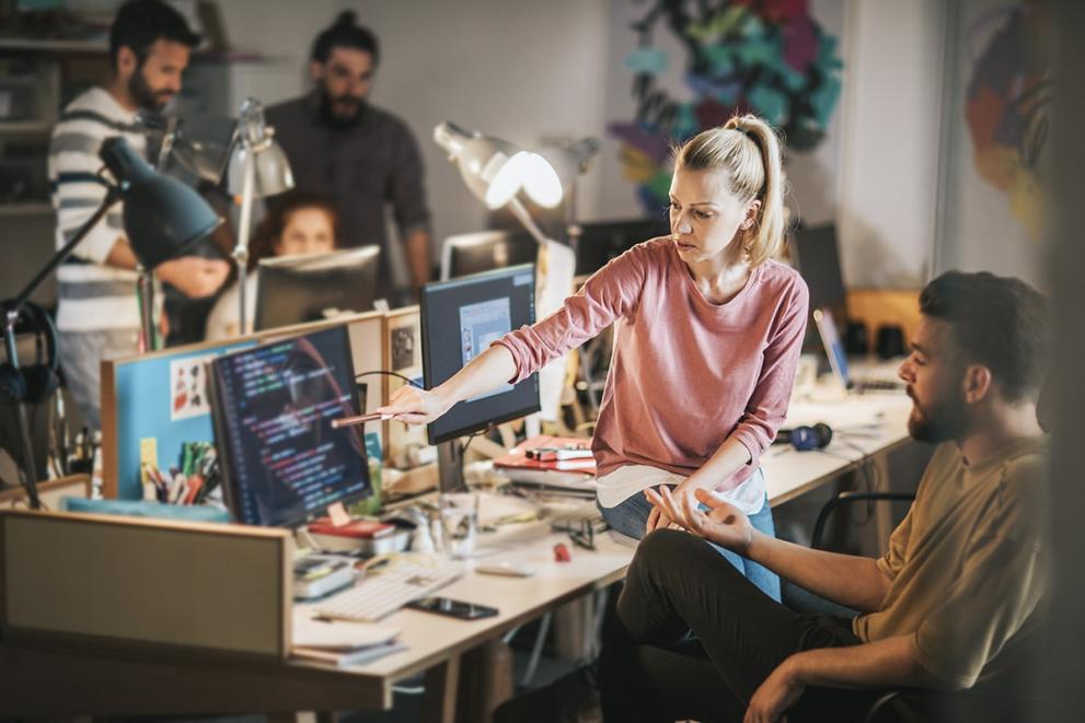 Two office workers in discussion in an office setting. One is seated on the edge of the desk and is pointing at information on the computer screen in front of them.