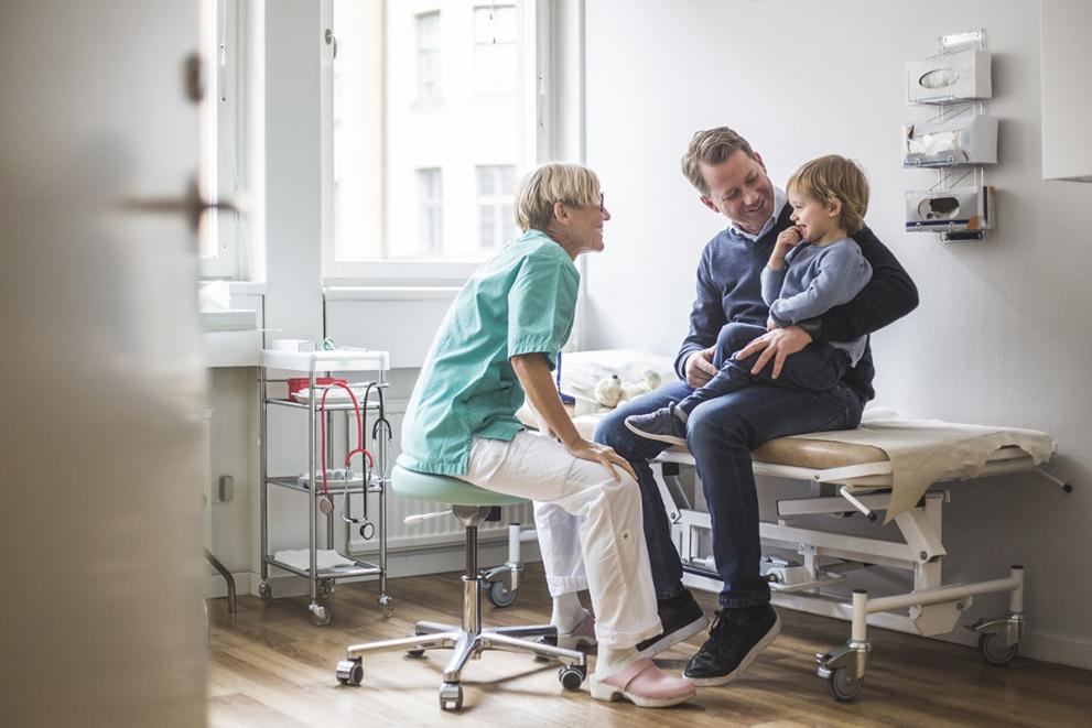 Meditsiinitöötaja vestleb mehega, kes hoiab väikest last süles. Asukohaks on haigla konsultatsioonituba.