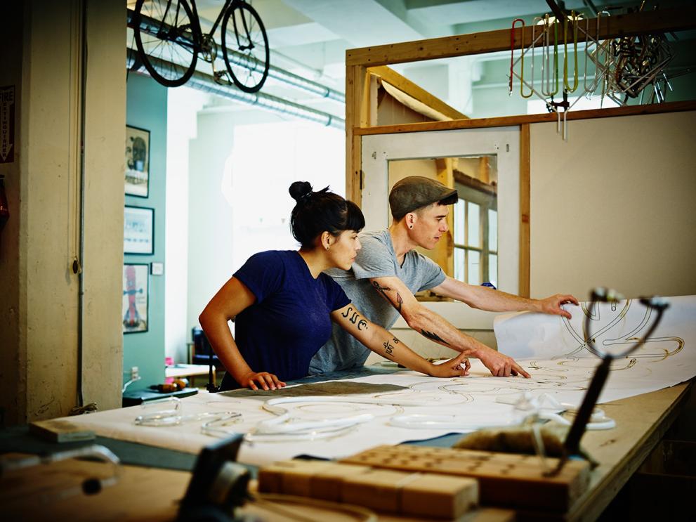 Two young workers in a workshop setting studying plans on large paper sheets spread out on a worktable in front of them.