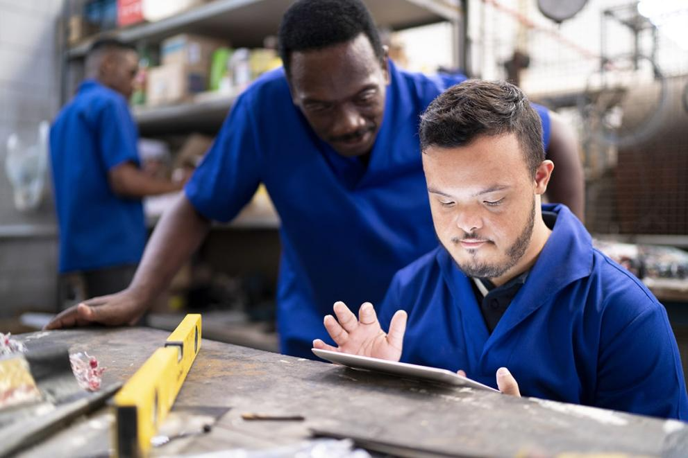 Two workers in blue work coats looking at a tablet in a factory setting. One is seated and holding the tablet while the other is standing behind and looking over his shoulder.